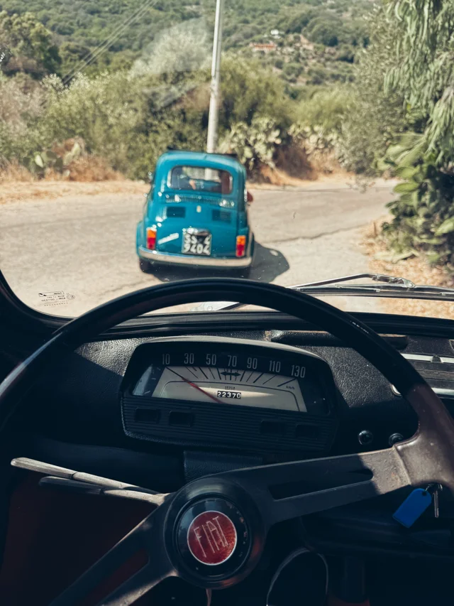 View from a Fiat 500 following a turquoise vintage Fiat along a Sardinian road